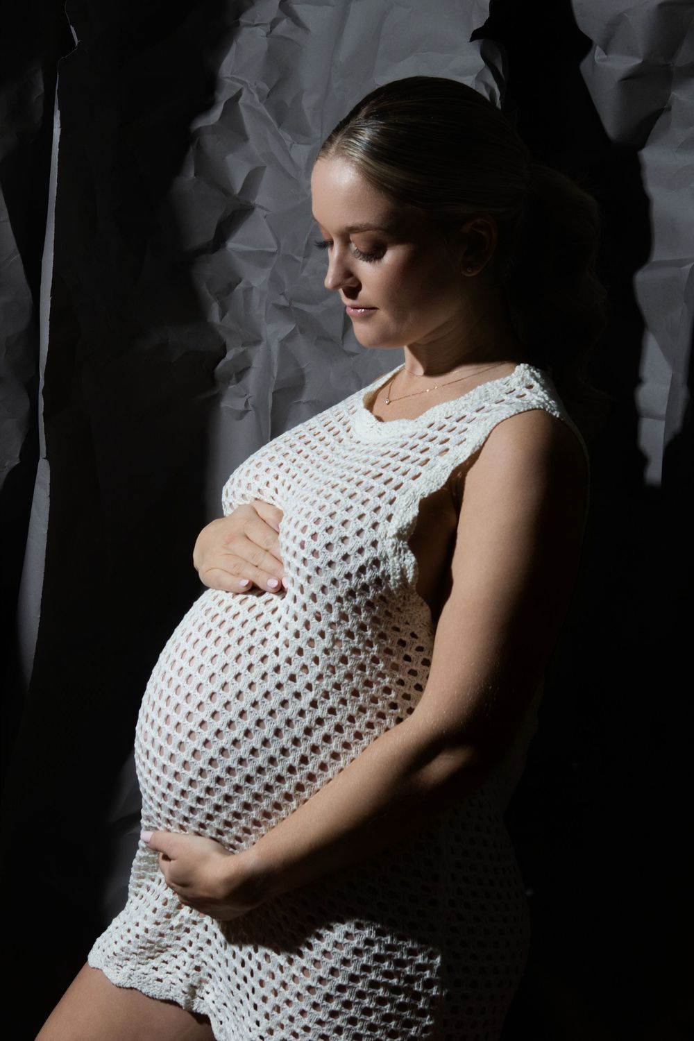 Pregnant woman in white crochet dress cradling her baby bump against a dark textured background.