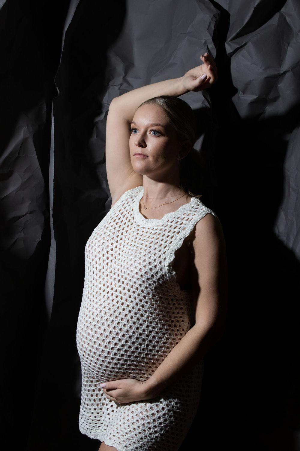 Pregnant woman in white crochet dress poses gracefully against dark crumpled background with arm raised.
