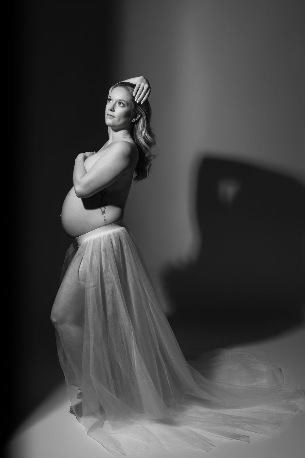Black and white maternity photo of a pregnant woman in a flowing sheer skirt, posing gracefully in a studio setting.