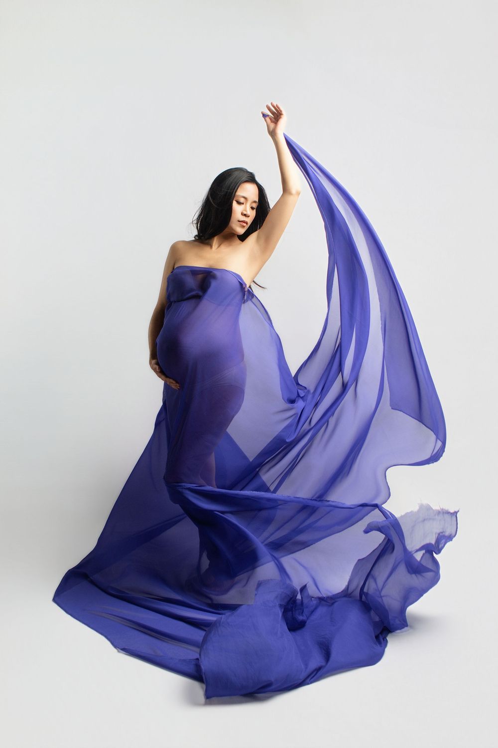Pregnant woman draped in flowing blue chiffon fabric, posing gracefully against a white background.