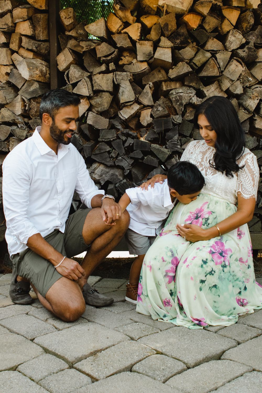 Family sharing a tender moment together in front of stacked firewood wearing white and floral outfits.