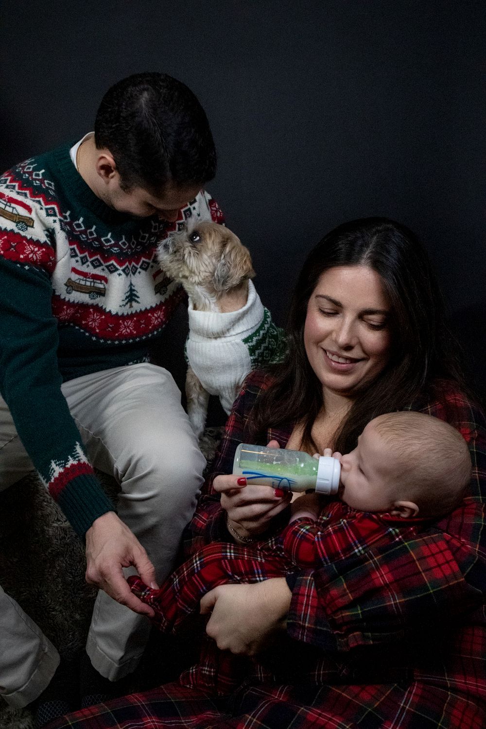 Family wearing holiday sweaters sits together with their dog and small baby during Christmas season portrait session.