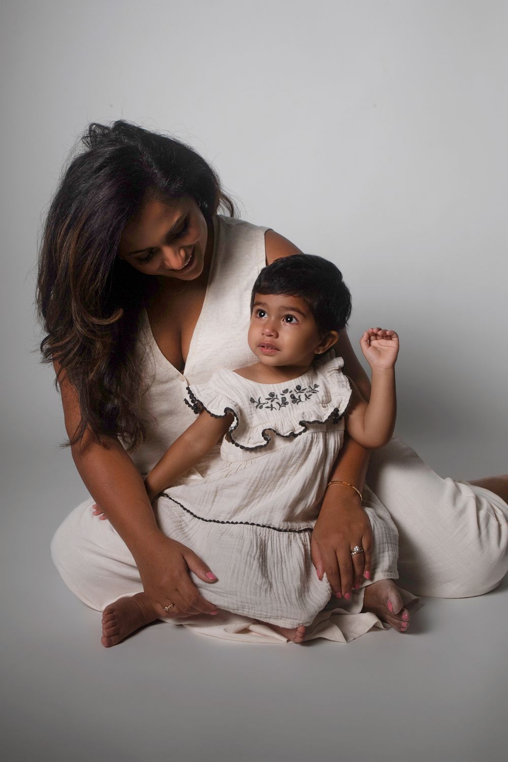 A mother and young child wearing matching white outfits pose together against a gray backdrop in a studio setting.