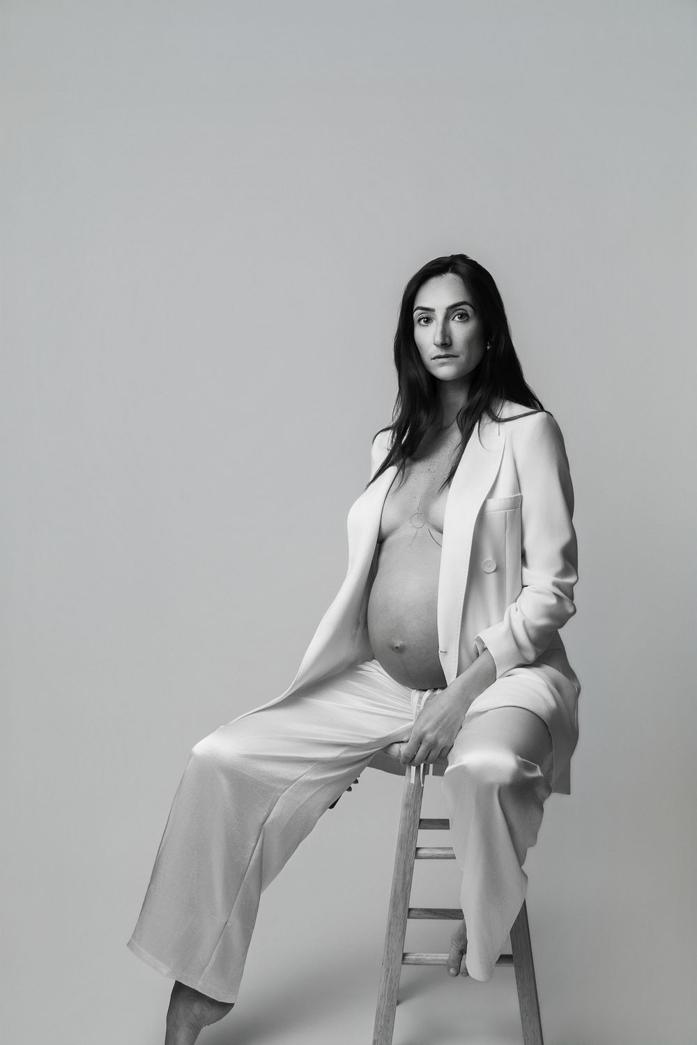 Black and white portrait of a pregnant woman in a white blazer and pants sitting on a stool.