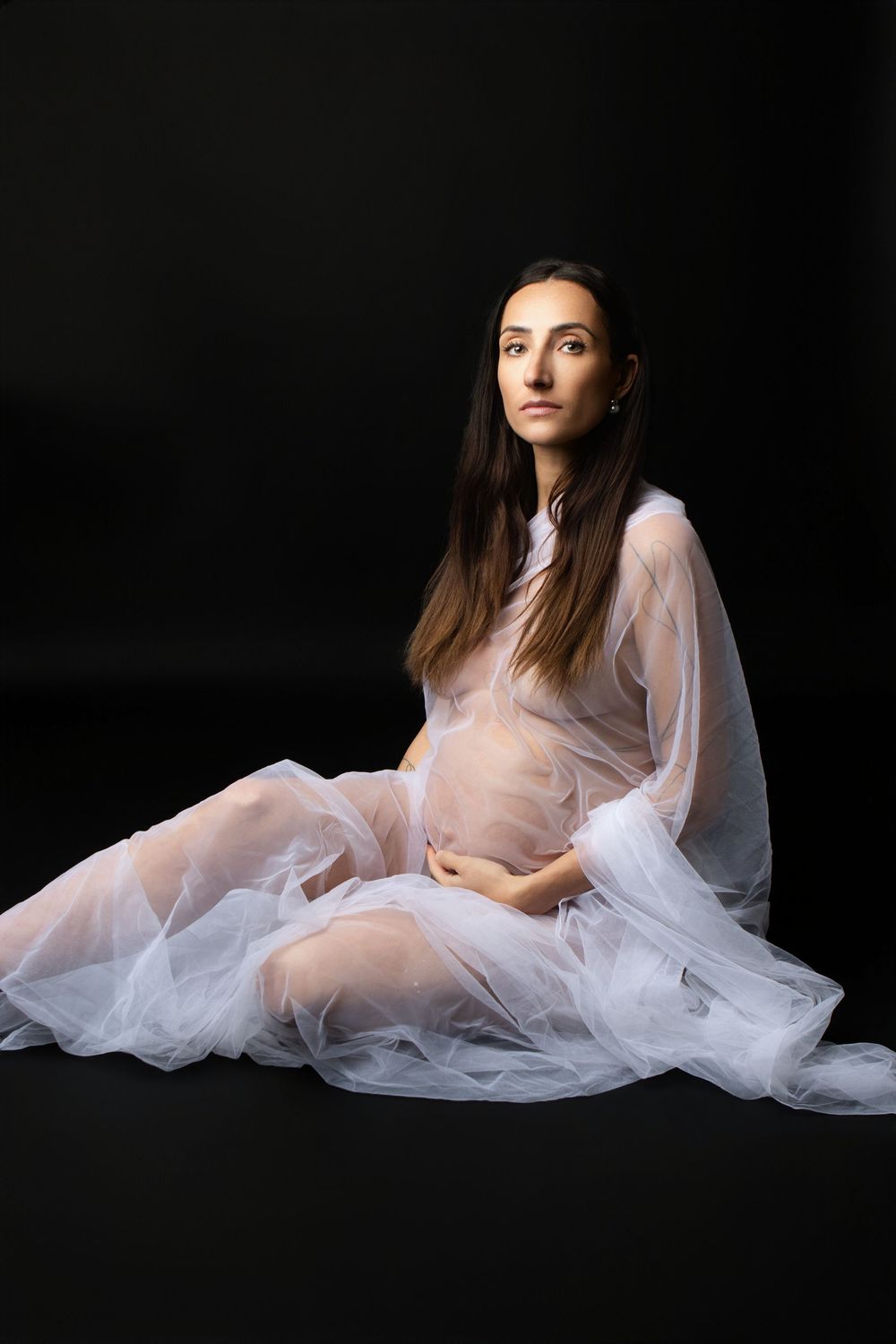 Pregnant woman in sheer white flowing gown sitting gracefully against black background, cradling baby bump.