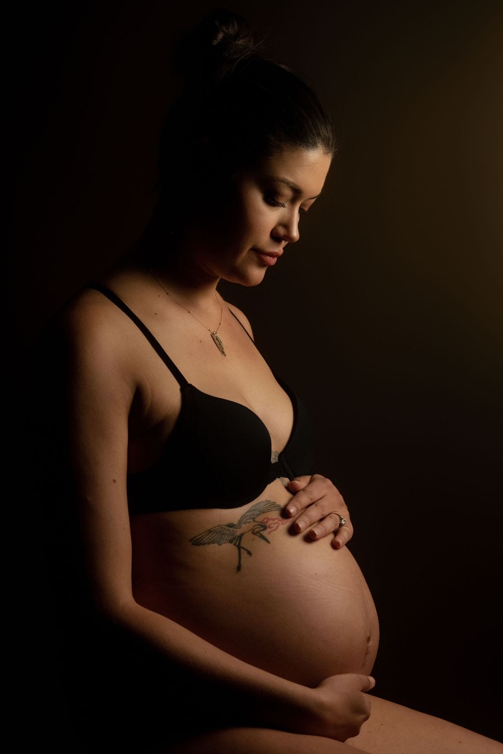 Artistic maternity photo in dark moody lighting shows expectant mother sitting in black bra with tattoo on her belly.