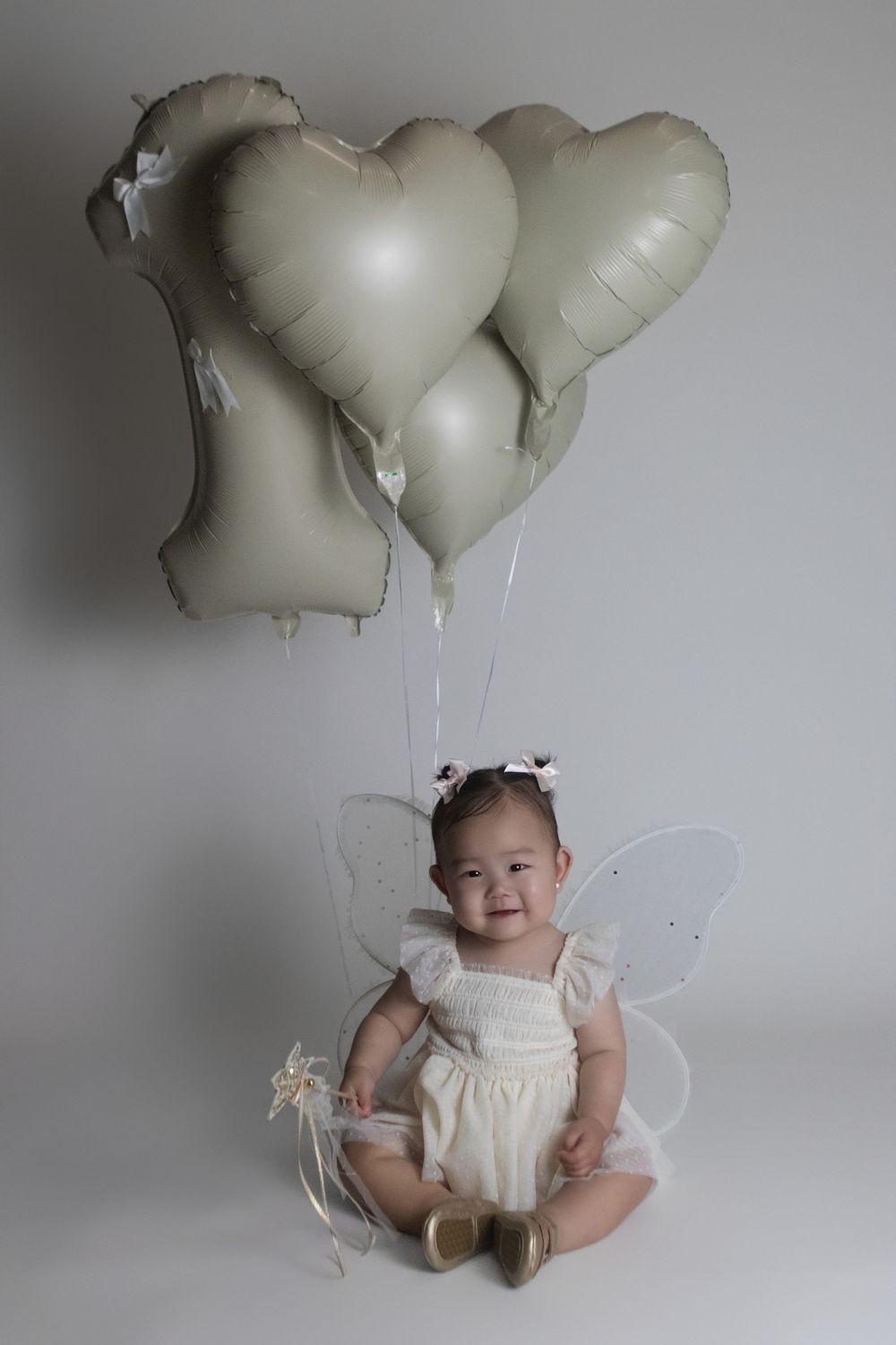 Small child wearing white fairy wings and dress sits on floor with three white heart-shaped balloons floating above.
