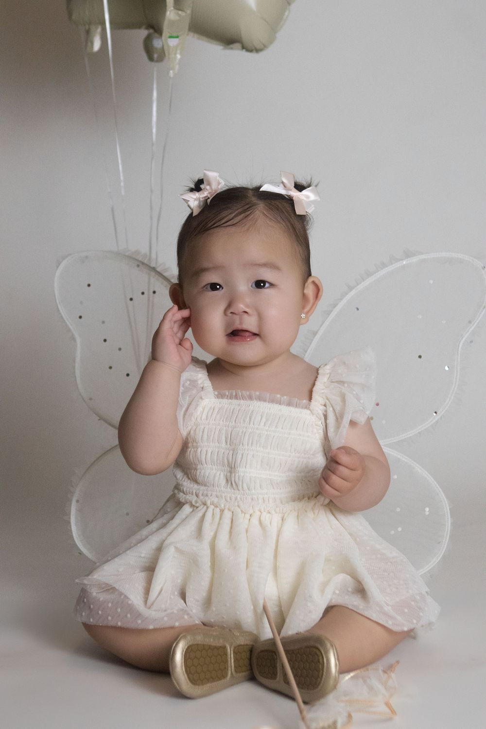 A baby wearing a cream-colored dress and fairy wings poses for milestone photos against a neutral background.