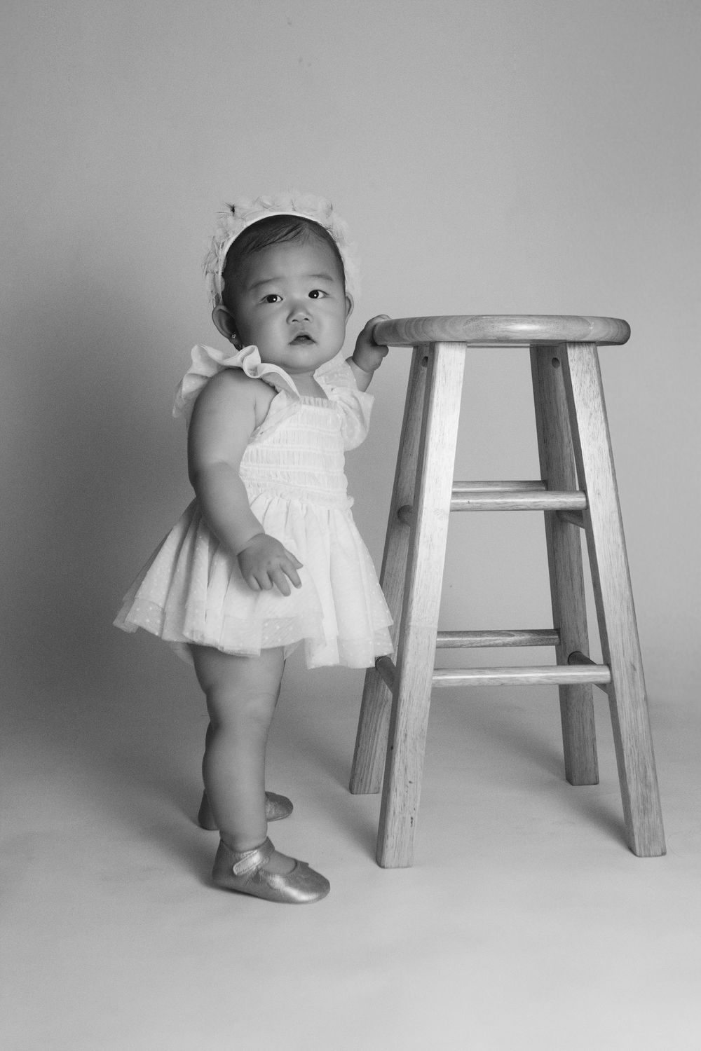 A series of black and white photos shows a baby in a white dress taking first steps while holding onto a wooden stool.