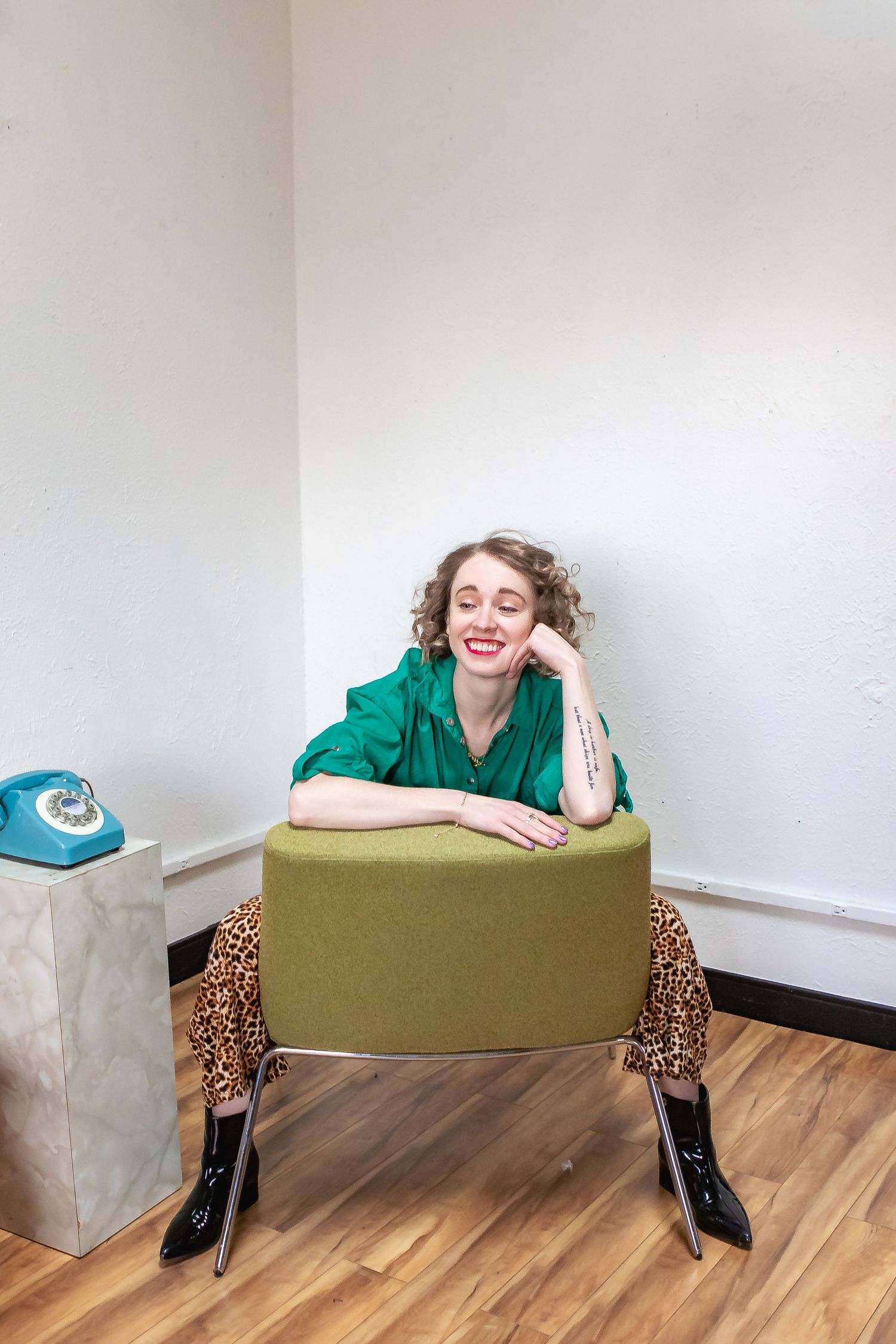 Person in green shirt and leopard pants poses playfully with vintage blue telephone and ottoman on hardwood floor.