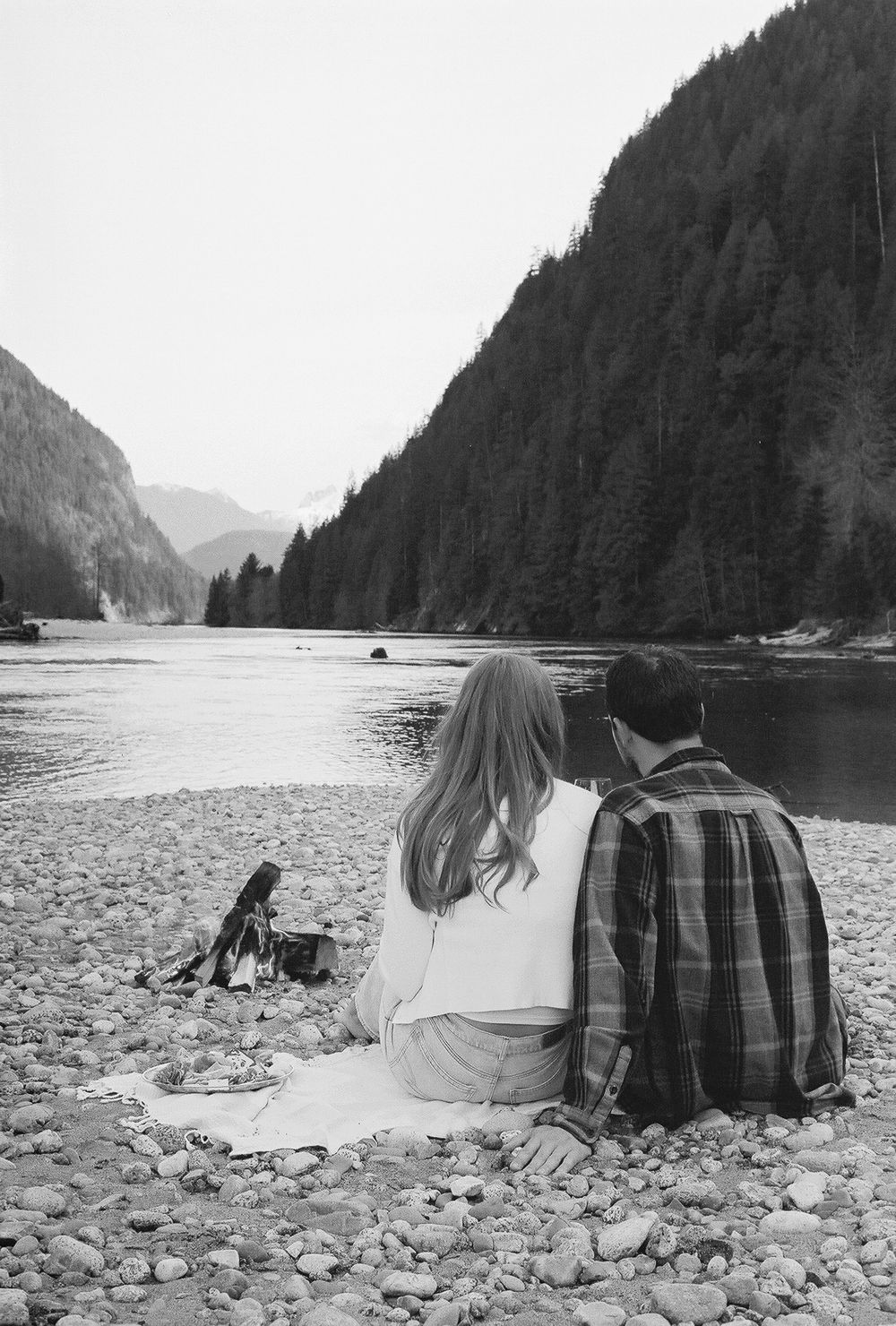 Couple sitting on rocky beach with mountain backdrop in black and white.
