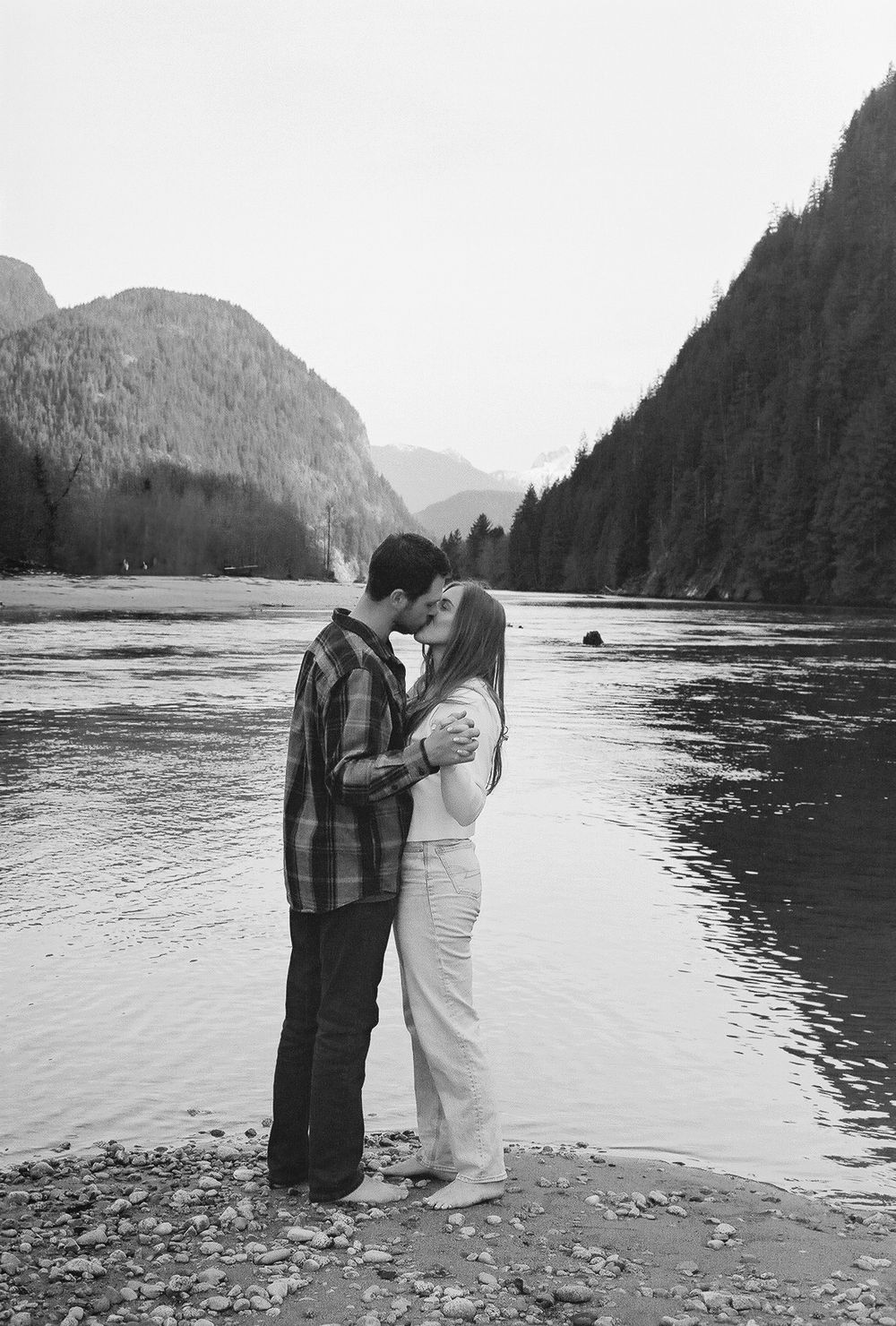 Couple standing together on rocky shore with mountainous lake backdrop.