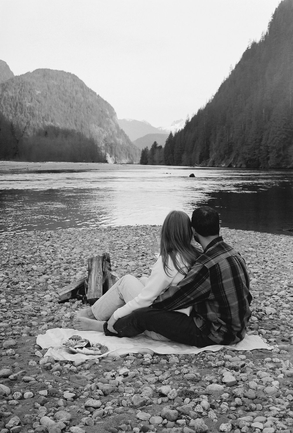 Couple lying on rocky beach shore with mountains and lake in background.