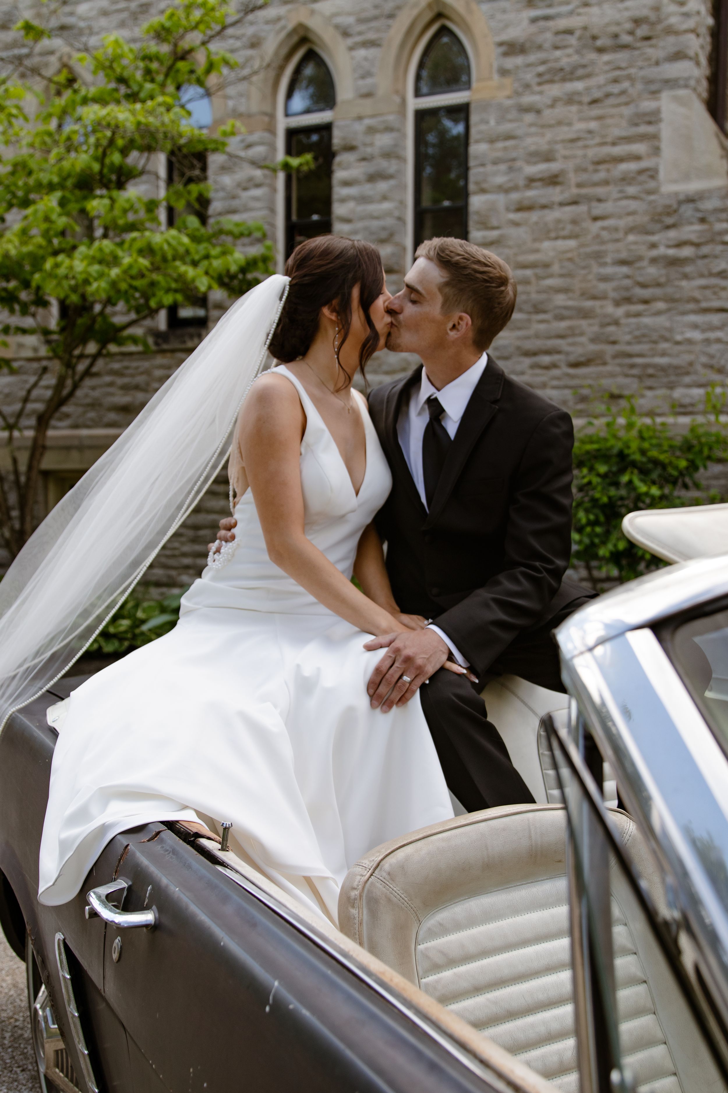 Newlyweds share romantic moment in classic car near stone church.