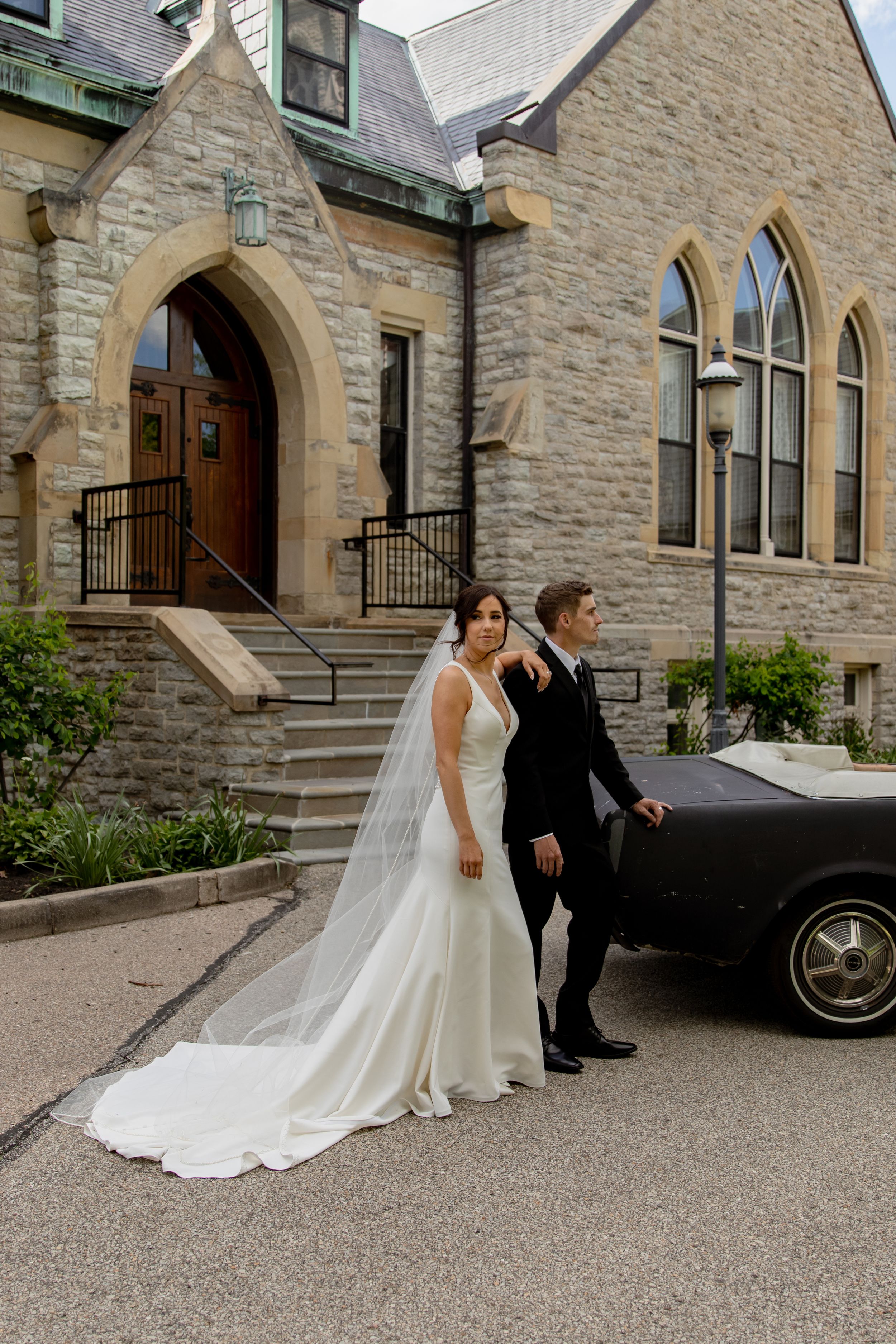 Wedding couple sits near vintage car outside stone church building.