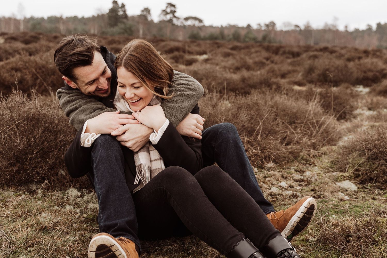A couple sit in a field of heather. Their arms are wrapped around each other and they are laughing.