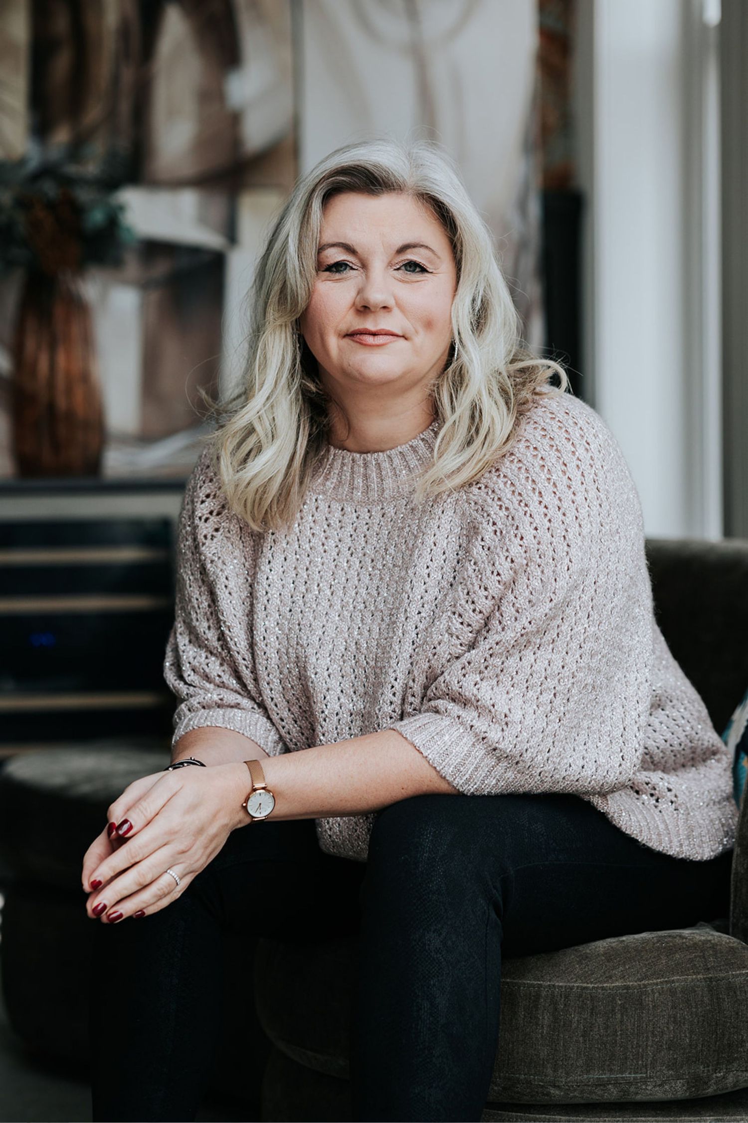 Person in cozy beige knit sweater sits casually on steps, styled with blonde wavy hair and dark pants in moody lighting.