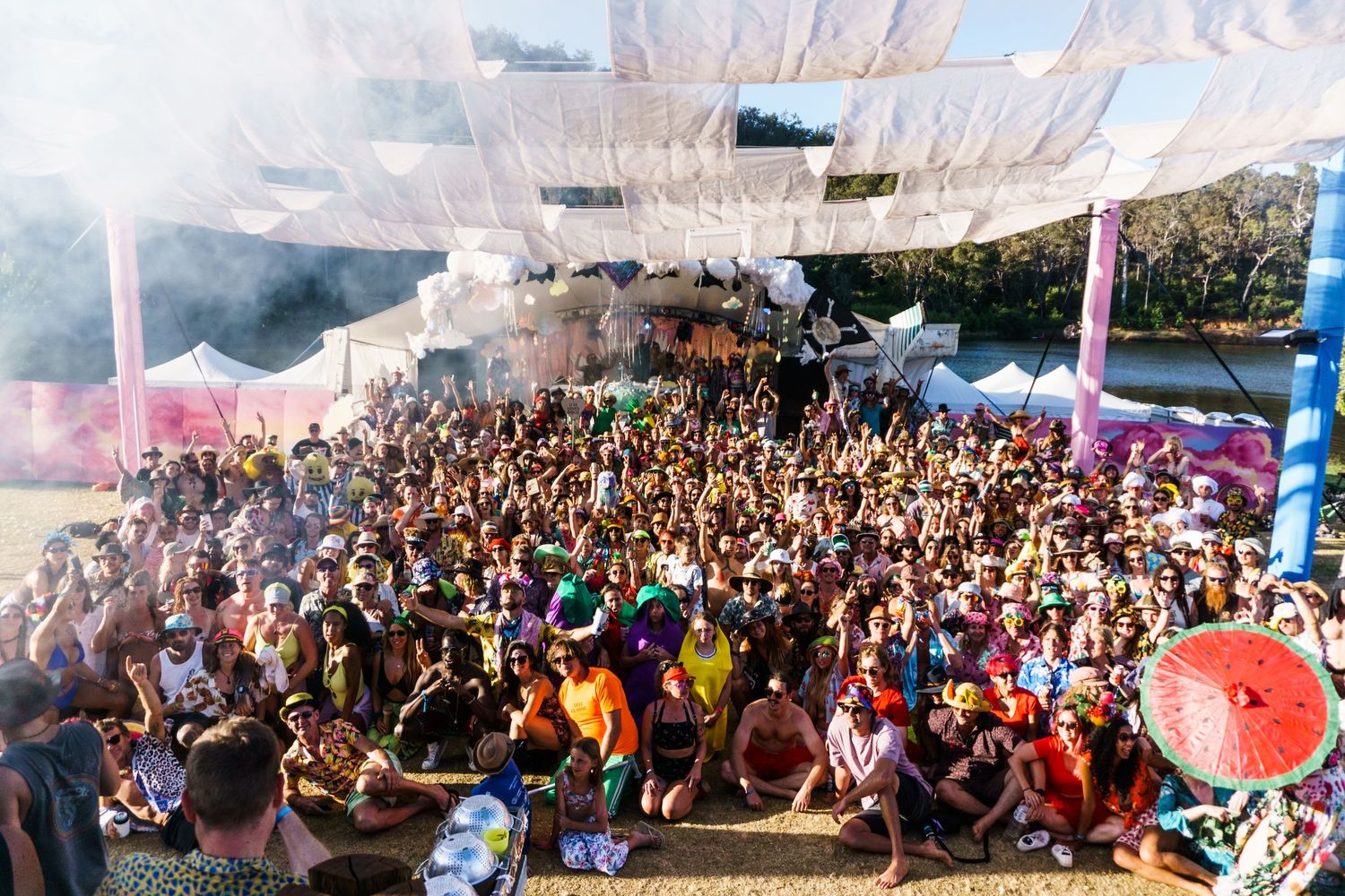 A large outdoor festival crowd gathered under colorful banners with smoke effects during a daytime event.
