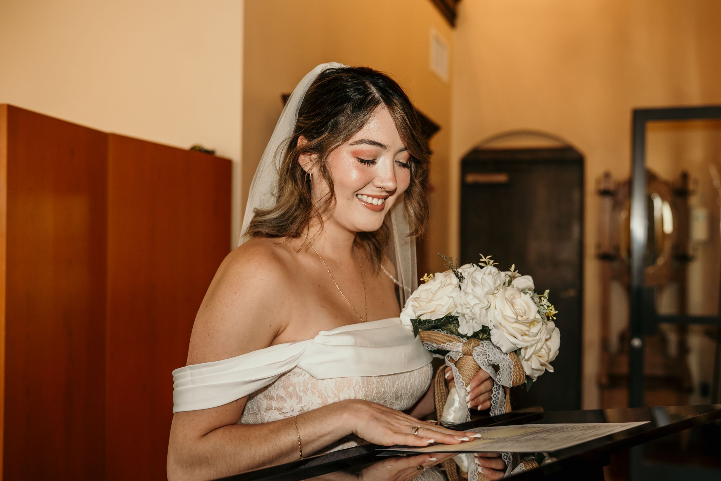 A person in an off-shoulder cream dress smiles while holding a bouquet of white roses indoors.