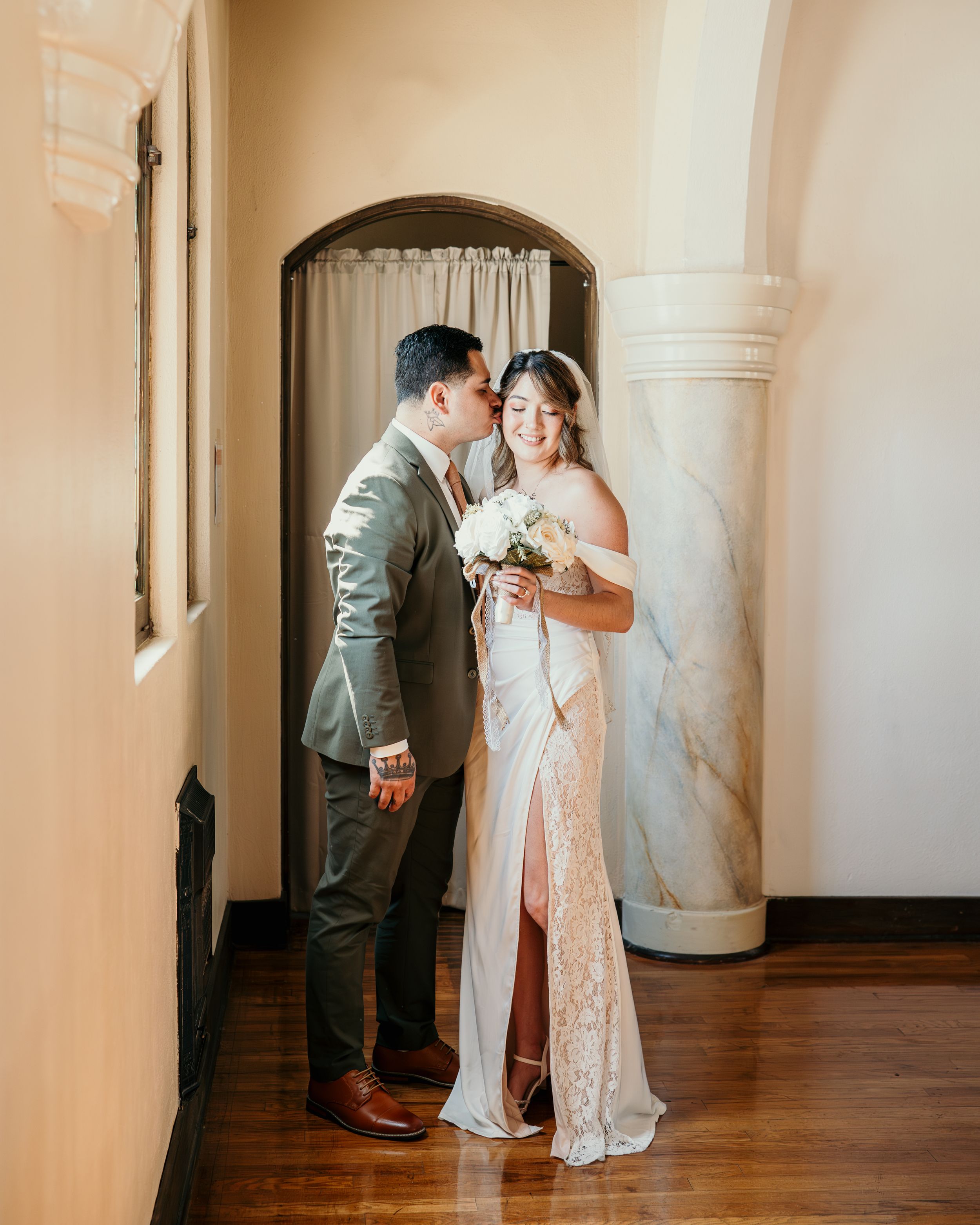 Newlywed couple shares an intimate moment in an elegant hallway with arched doorways and sunlit cream walls.