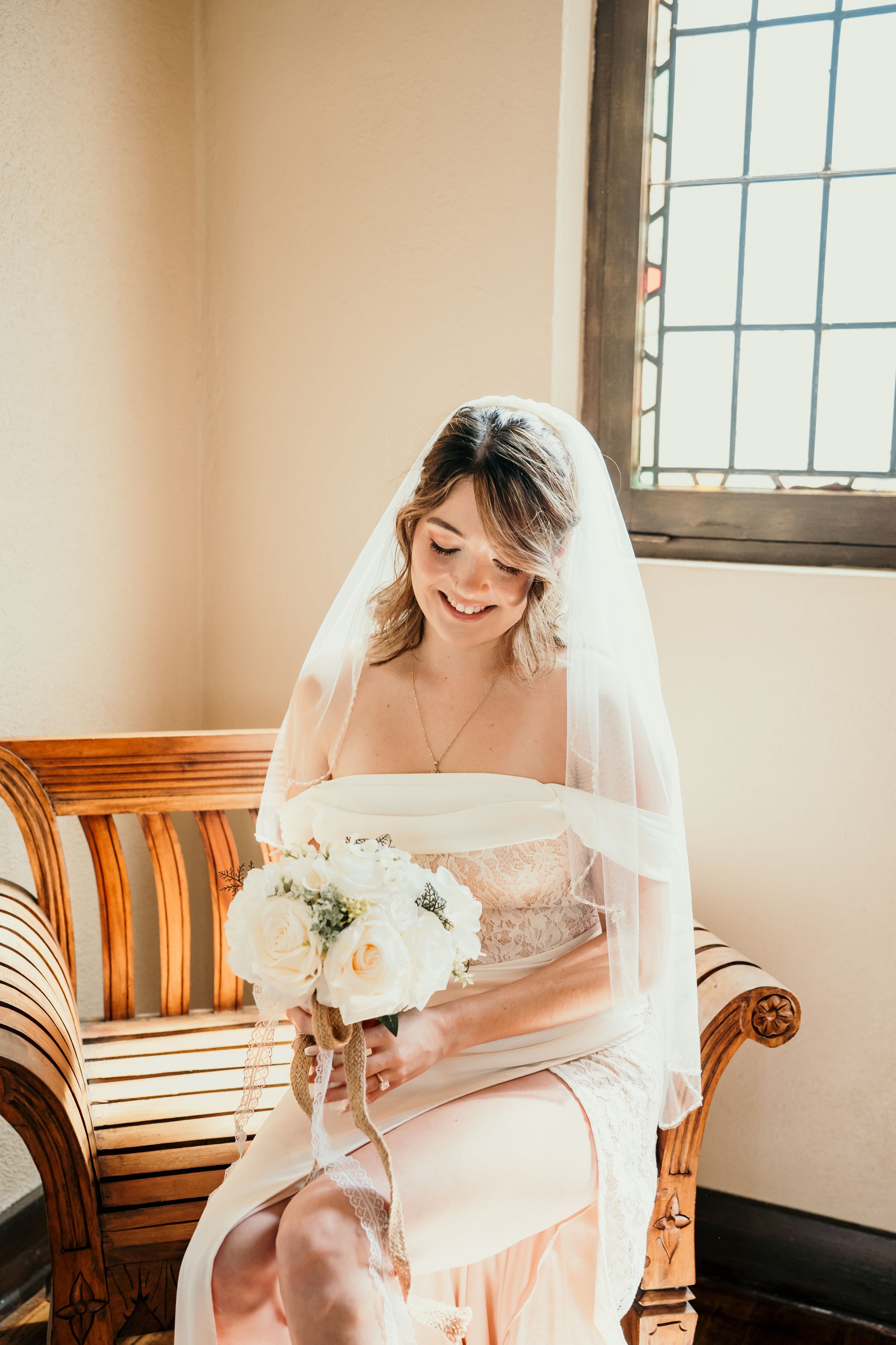 A bride in a white dress and veil sits on a wooden bench holding a bouquet of white flowers by a window.
