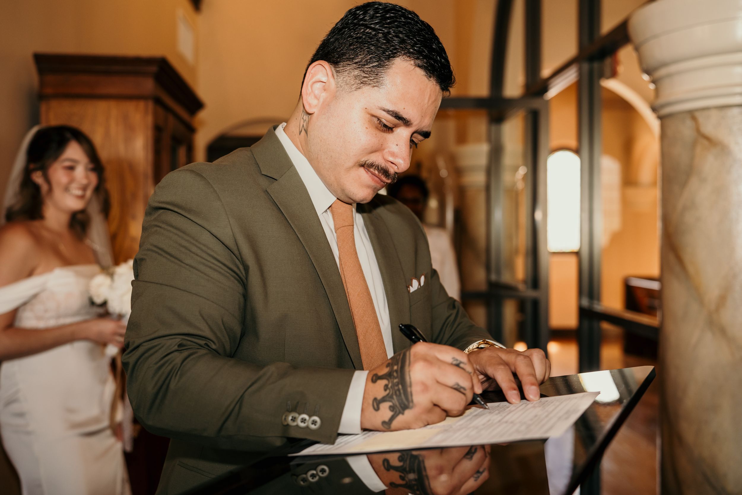 Guest signs wedding document while standing in elegant venue hallway wearing brown suit and orange tie.