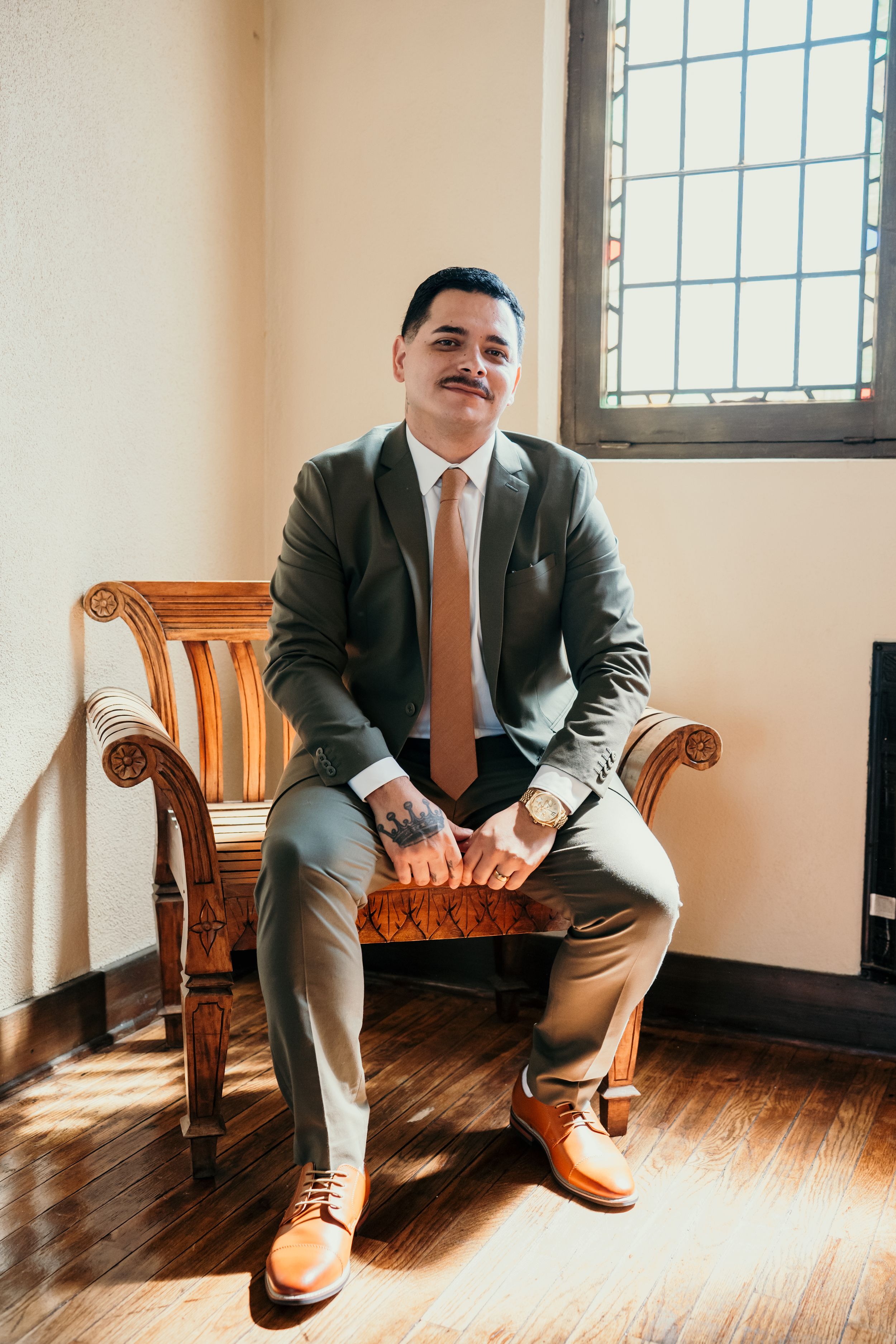 Professional dressed in a grey suit with tan shoes sits on a wooden armchair by a window with natural light.