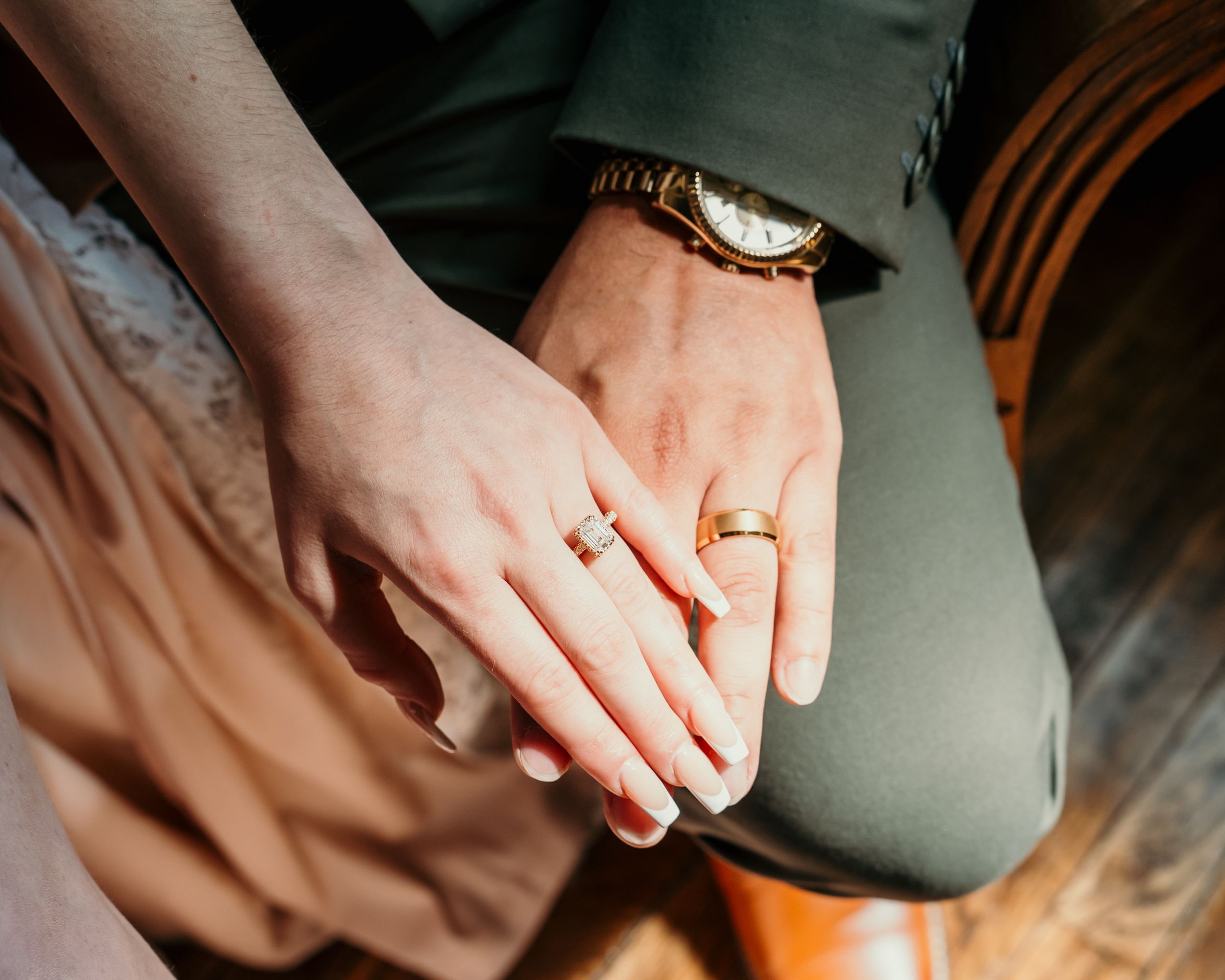 Hands clasped together showing wedding bands and red nail polish against a green and peach background.