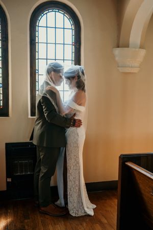 A couple shares an intimate moment by a church window, with the bride wearing a flowing lace gown and groom in a gray suit.