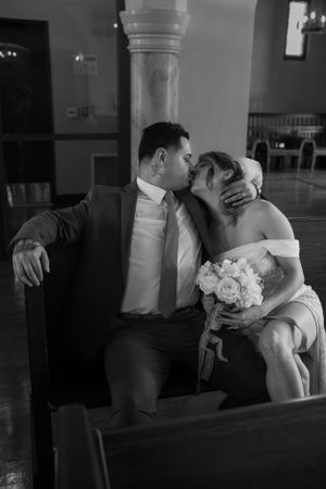 A tender moment captured in black and white as a couple in wedding attire share a romantic kiss on a church pew.