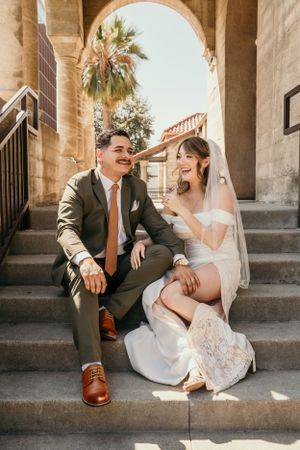 A joyful couple shares a candid wedding moment sitting on stone steps under an arched passageway with palm trees.