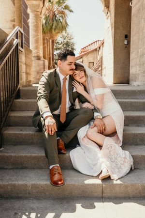 Couple in wedding attire sitting intimately on stone steps in a Mediterranean-style setting with palm trees.