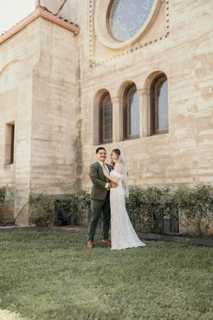 Wedding couple posing together in front of a historic stone church with arched windows.
