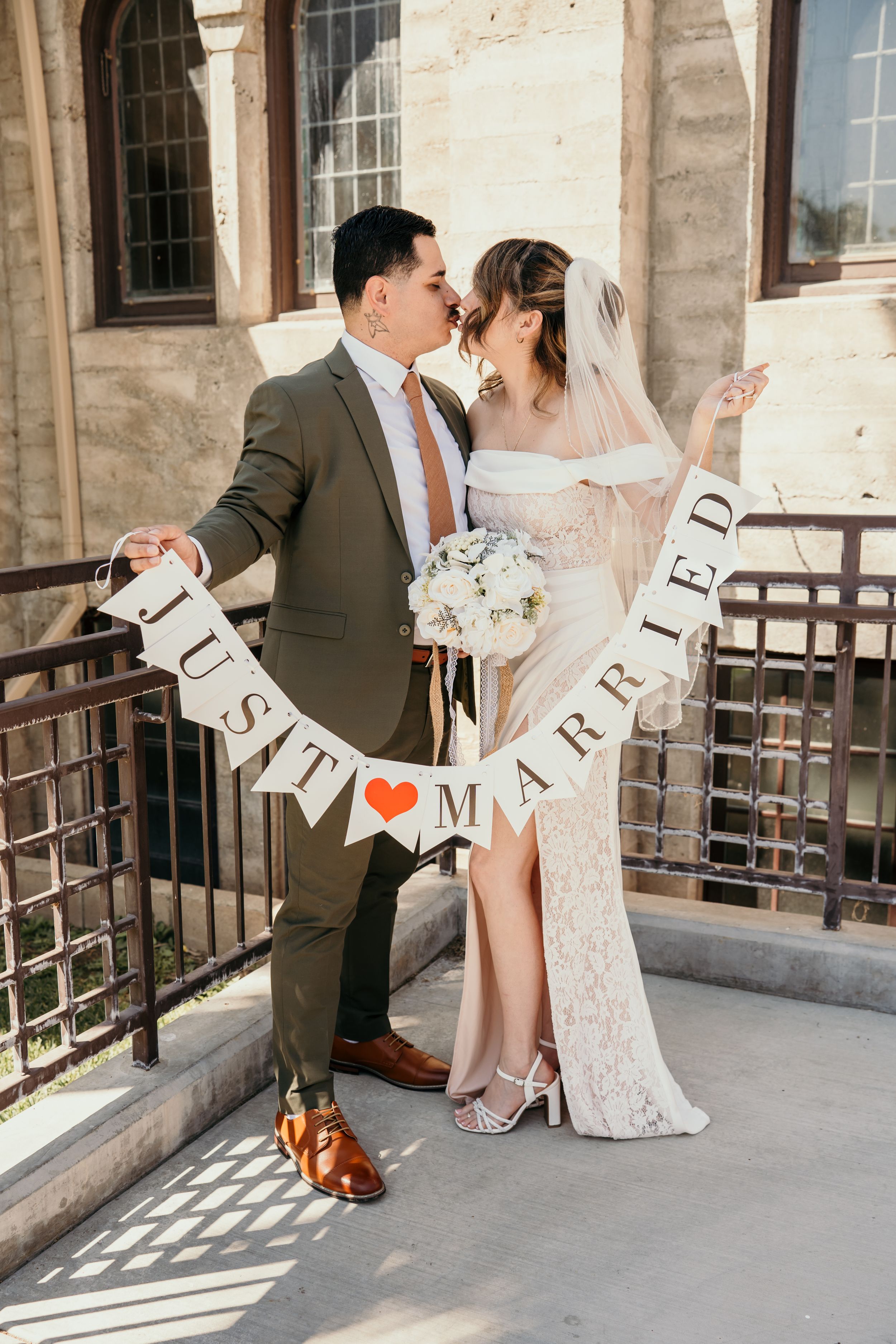 Newlywed couple poses with 'Just Married' banner at church venue, bride in lace gown holding white bouquet.