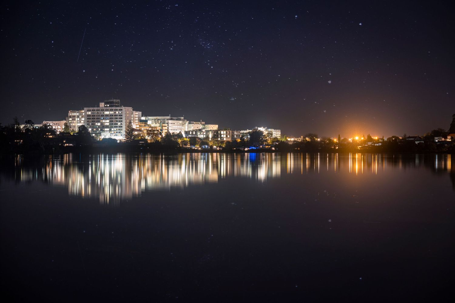 City lights reflected on calm water under a starry sky, with illuminated buildings lining the opposite shore.