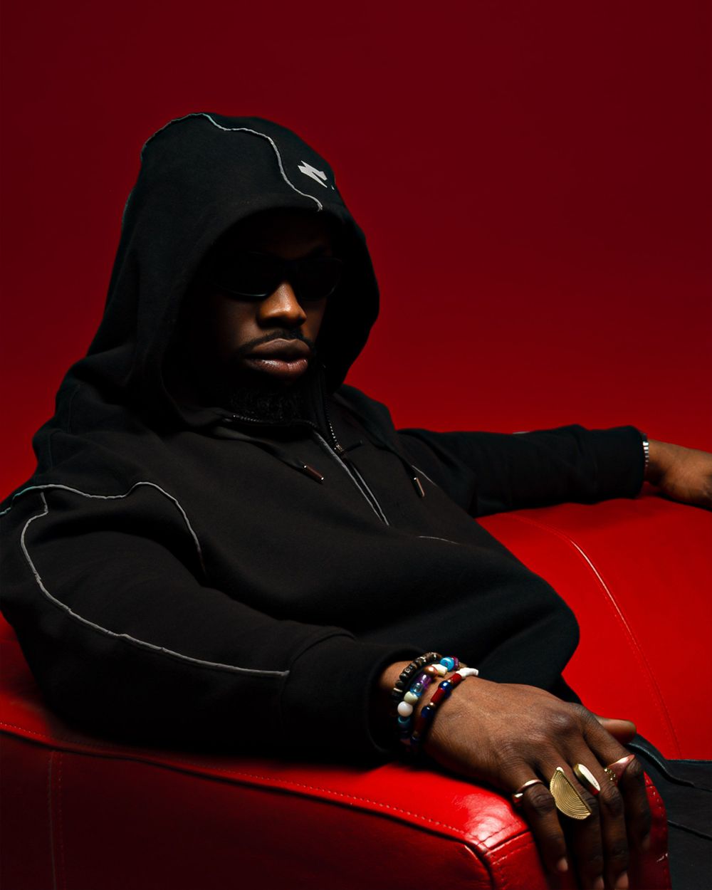 A person in a black hooded sweatshirt sits on a red leather chair against a dark red background.