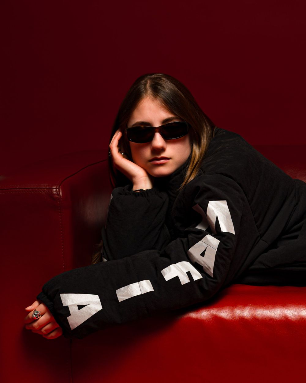 A person in sunglasses and a black sweater with text poses dramatically on a red couch against a dark red background.