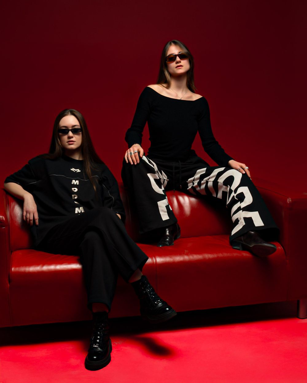 Two people in black outfits and sunglasses pose stylishly on a red couch against a deep red background.