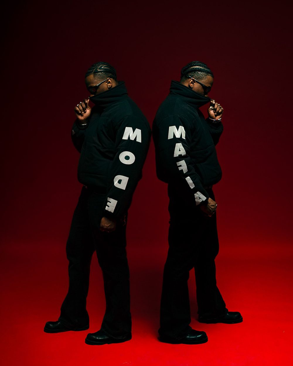 Two people stand back to back wearing black hoodies with MODE MALT text while posing against a dark red backdrop.