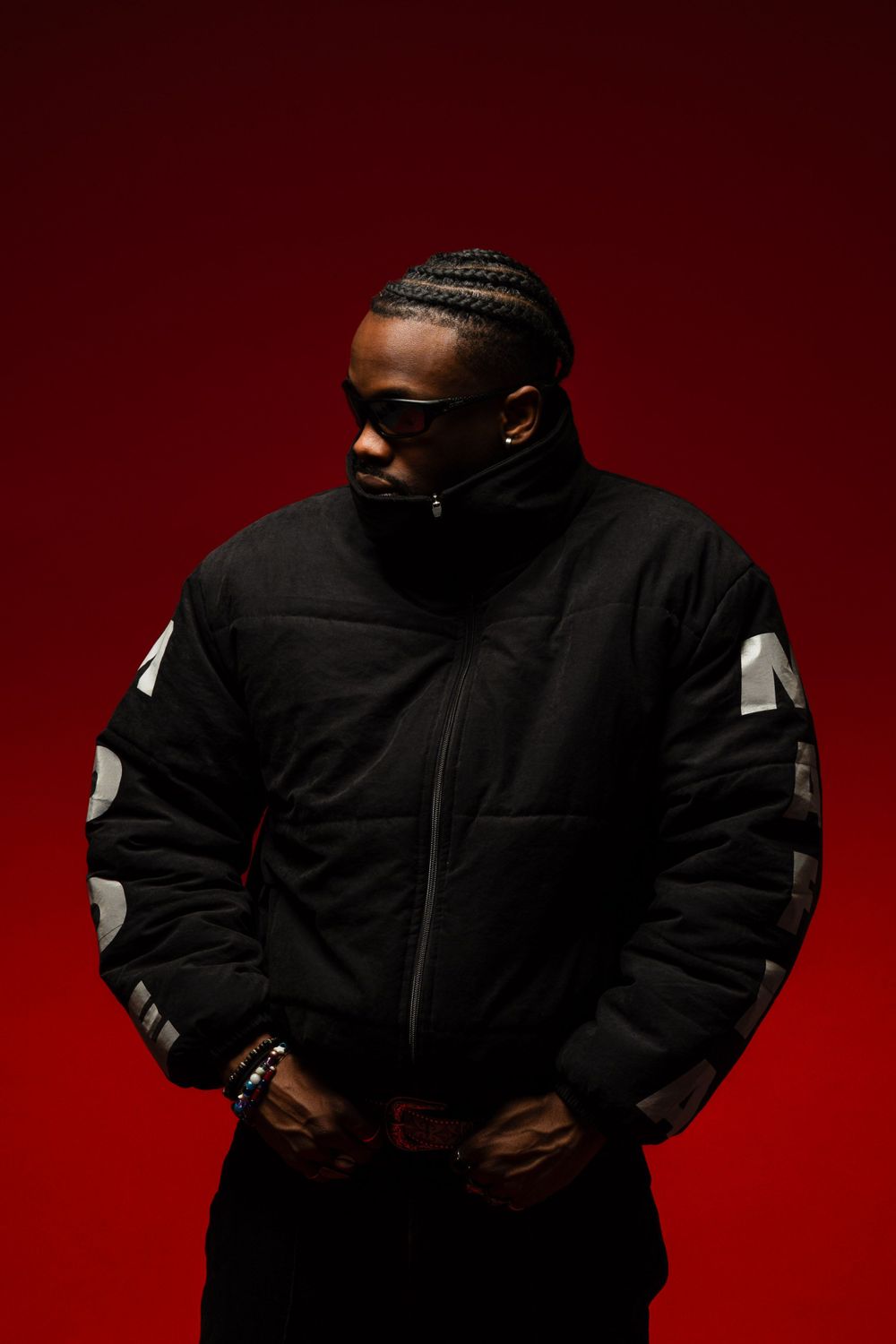 Stylish figure in black puffy jacket poses against dramatic red background in moody portrait.