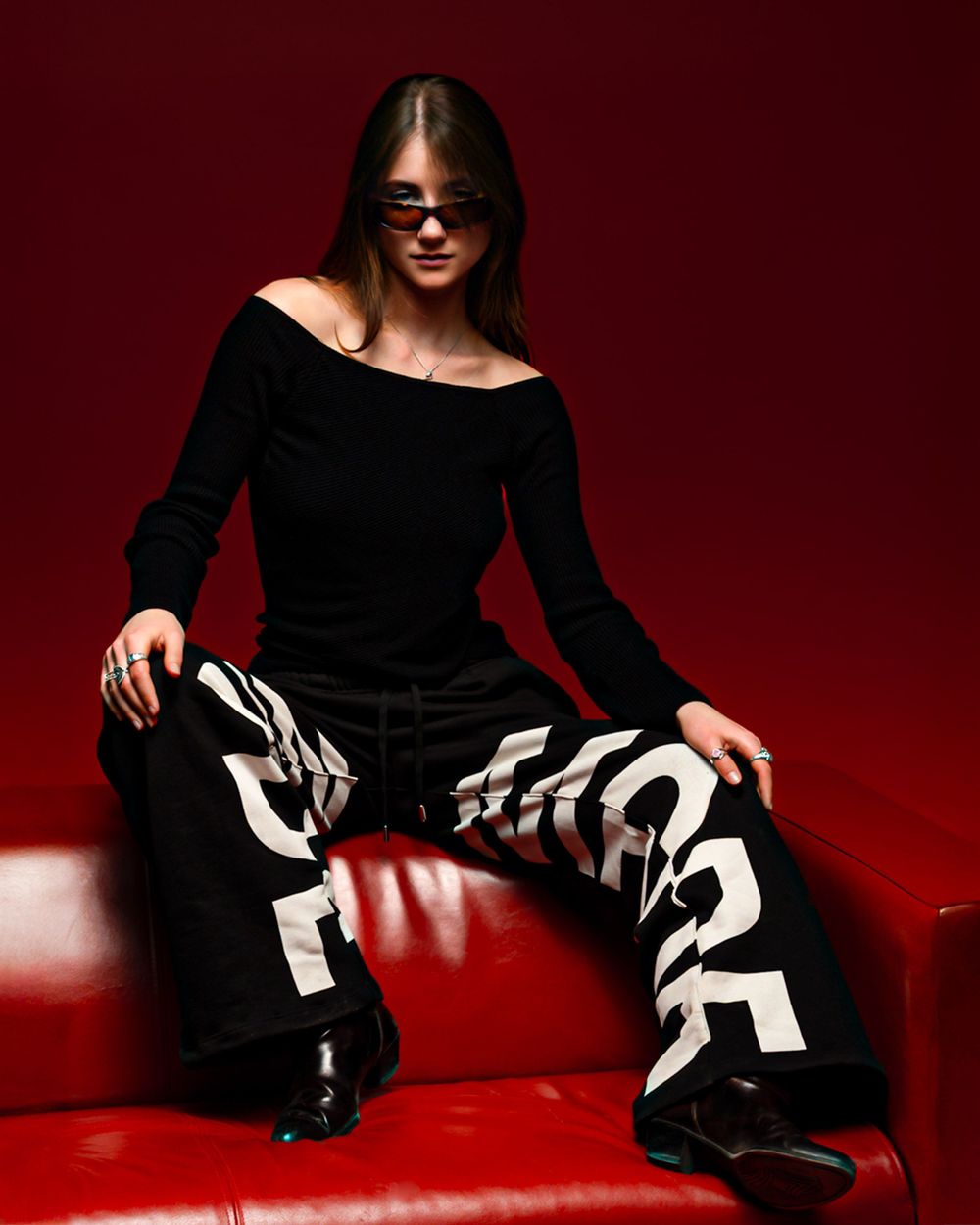 Model in black off-shoulder dress and striped pants poses on red leather couch against dark red backdrop.