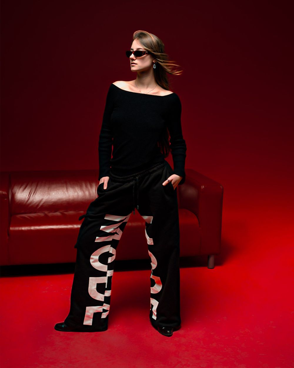 Model in black outfit with statement text pants poses dramatically on red leather couch against dark red backdrop.