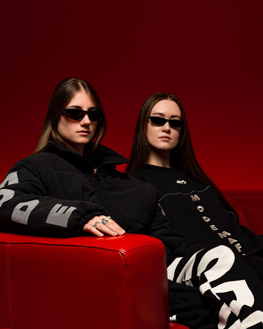 Two models in black streetwear and slim sunglasses pose on red leather seating against a dark red backdrop.