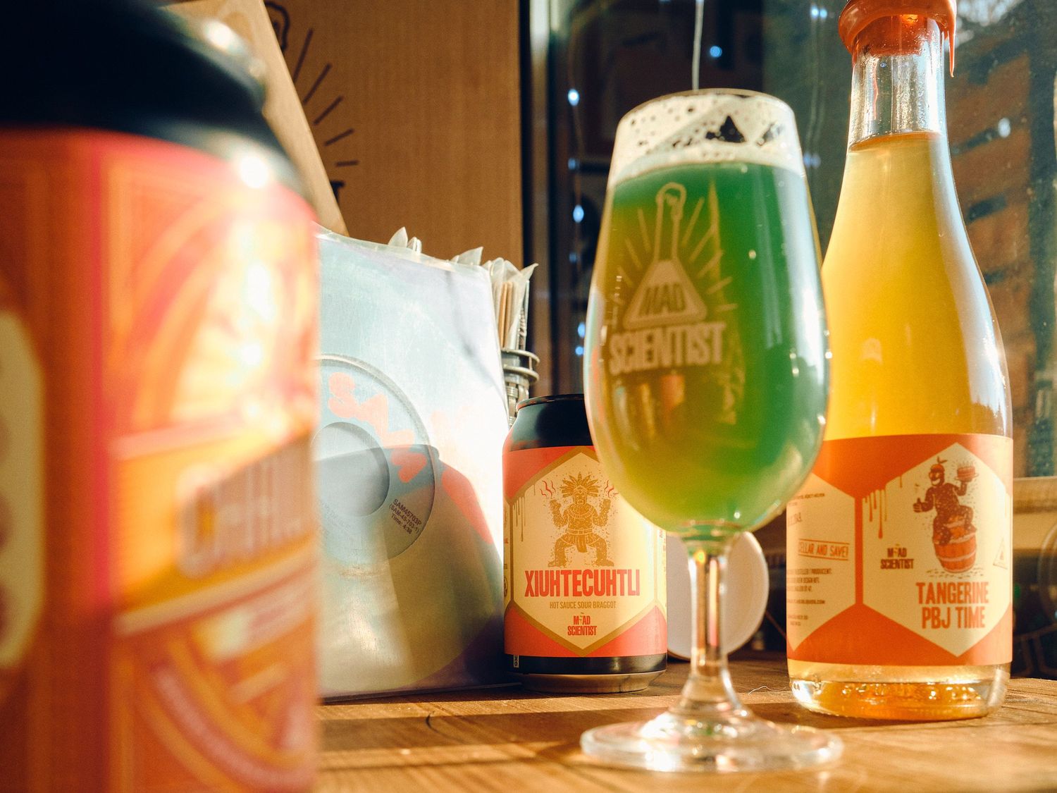 Colorful craft beer bottles and a stemmed glass filled with bright green beer displayed on wooden table in warm lighting.
