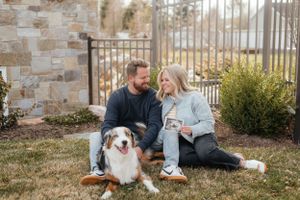 Couple sitting on grass with Australian Shepherd dog, holding ultrasound photo, smiling at each other near stone wall. Megan Snell Photo