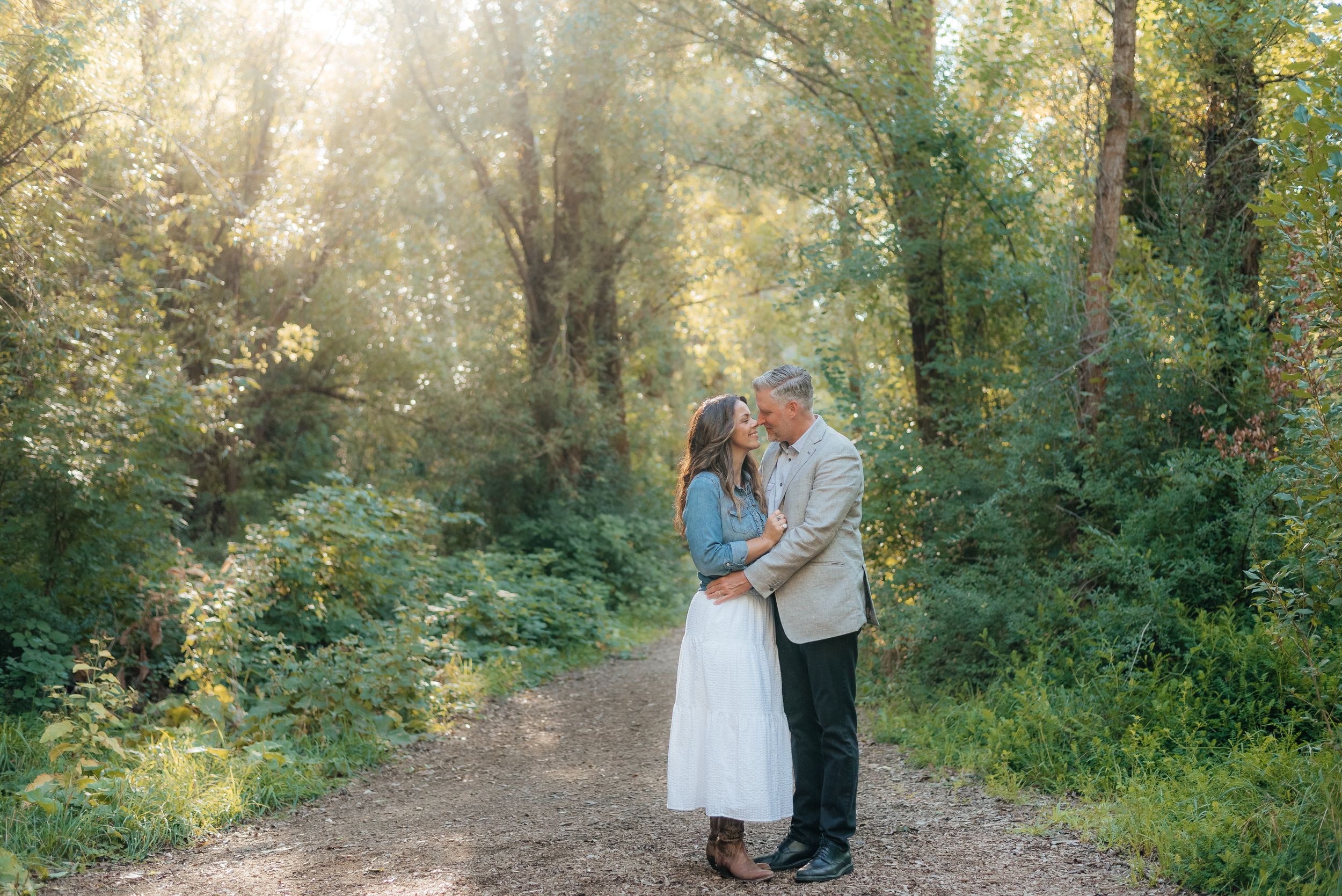 Couple sharing a romantic moment on a sunlit forest path surrounded by lush green trees and foliage.