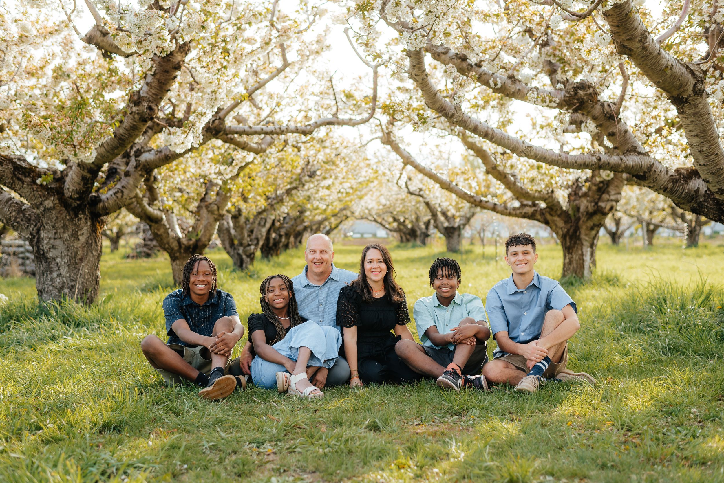 Family of six smiling together in a blooming orchard, sitting on green grass surrounded by flowering trees. Megan Snell Photo Utah Weddings