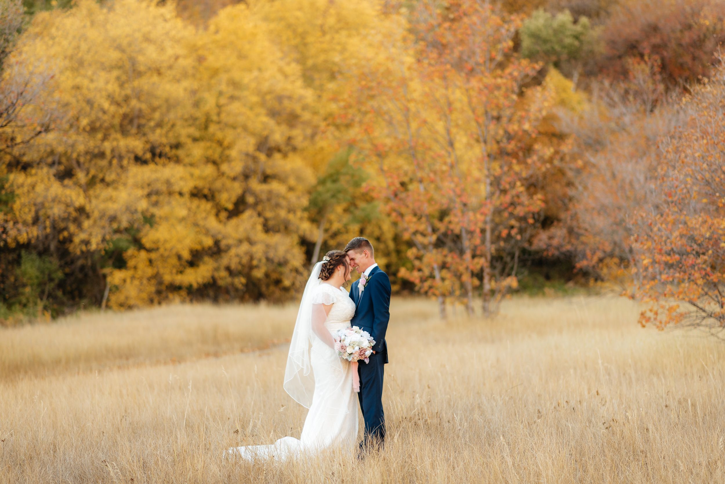 Bride and groom share a romantic moment in a golden autumn field surrounded by vibrant fall foliage. Megan Snell Photo Utah Weddings