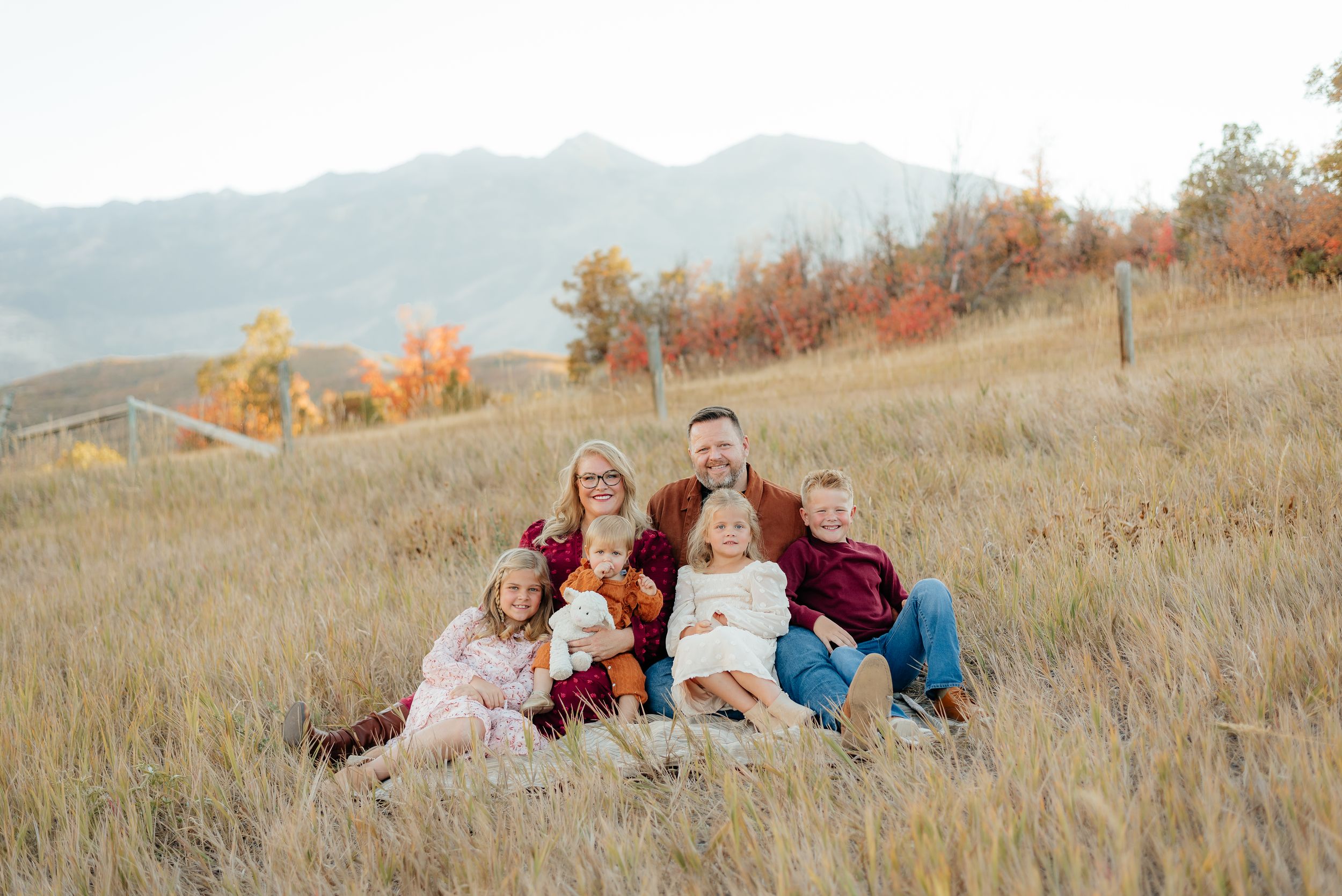 Family of six poses in golden autumn field with mountain backdrop and colorful fall foliage. Megan Snell Photo Utah Weddings