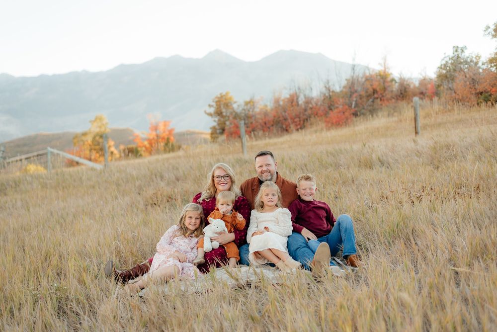 Family of six poses in golden autumn field with mountain backdrop and colorful fall foliage. Megan Snell Photo Utah Weddings